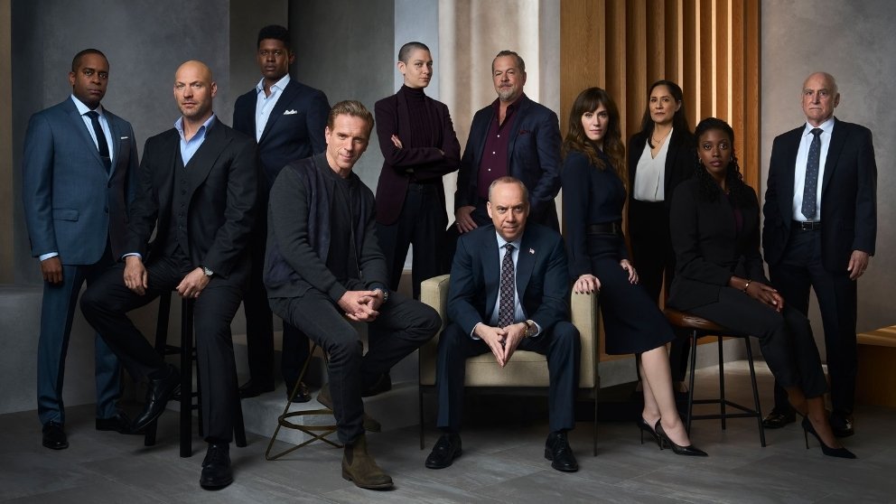 The main cast of the TV show Billions poses in a formal setting. Some are seated, while others stand in the background in business attire.