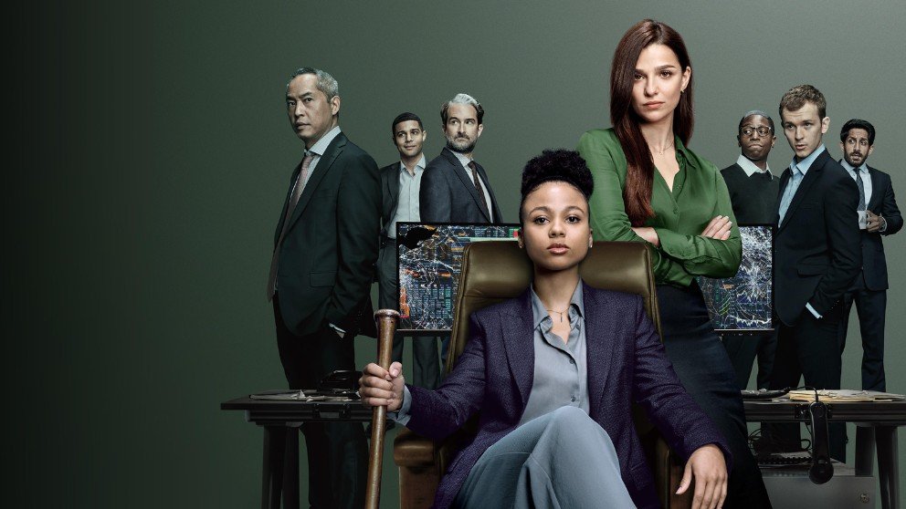 The main cast of the TV show Industry poses in a corporate setting. A woman in a suit sits in a chair holding a bat, while others stand behind her with financial data on screens in the background.