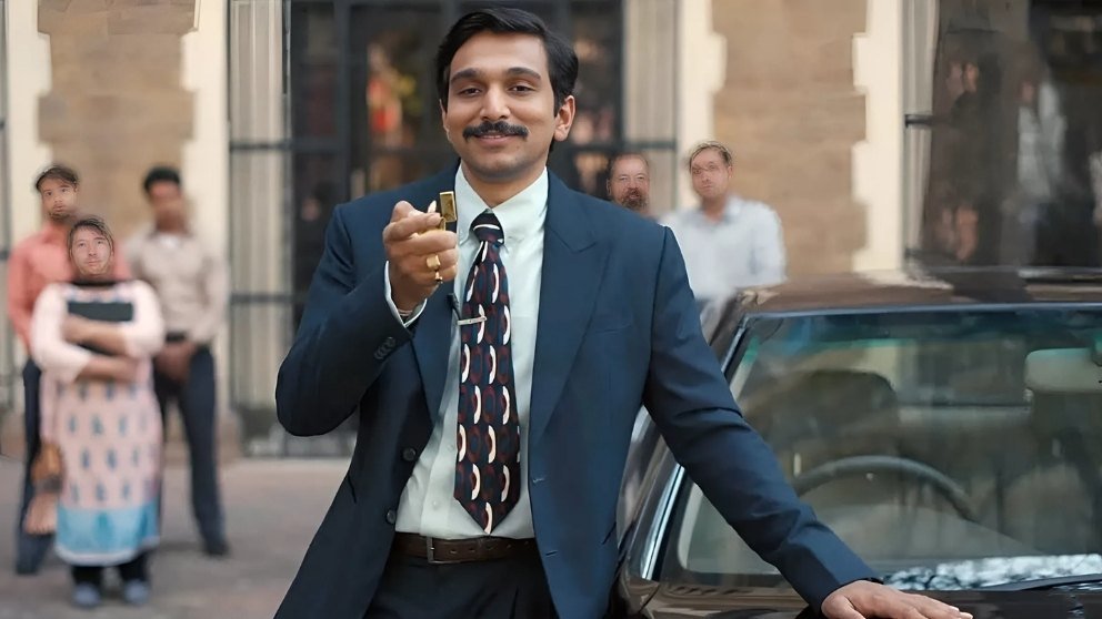 Pratik Gandhi as Harshad Mehta in Scam 1992, wearing a suit and holding a lighter, leaning on a car with people in the background.