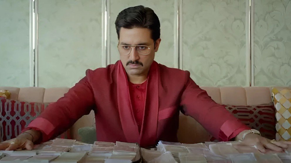 Image from The Big Bull showing Abhishek Bachchan in a red suit, sitting at a table stacked with bundles of cash, looking serious.